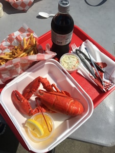 A culinary scene featuring a red tray with a container of freshly cooked lobster accompanied by a small cup of lemon butter and a slice of lemon. To the left, there is a basket of golden French fries in checkered paper. A bottle of Diet Coke, a small cup of coleslaw, and dining utensils including a fork, knife, and lobster crackers are also visible. The setting suggests a casual dining experience, possibly outdoors.
