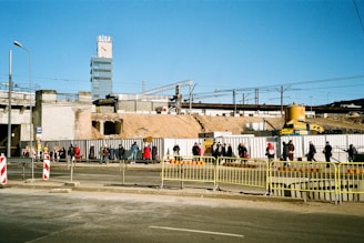 A construction site with people walking along a fenced-off area. In the background, there are cranes, a partially constructed building, and a tall building with the word 'RIGA' on top. The scene is set outdoors under a clear blue sky, with scattered pedestrians visible near barriers and temporary fencing.