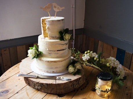 A two-tiered rustic wedding cake with a semi-naked frosting presentation is decorated with white roses and leaves. The cake sits on a wooden slab and is topped with a 'I Do, Me Too' bird design. Nearby are white flowers and a jar with string lights, creating a cozy and romantic atmosphere.