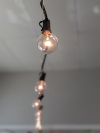 A series of round light bulbs hanging from a black cable, arranged in a line. The closest bulb is in focus, showcasing intricate filament details, while the others are progressively blurred, creating a depth effect against a neutral gray background.