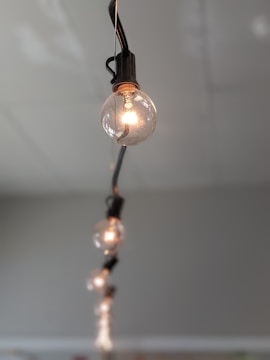 A series of round light bulbs hanging from a black cable, arranged in a line. The closest bulb is in focus, showcasing intricate filament details, while the others are progressively blurred, creating a depth effect against a neutral gray background.