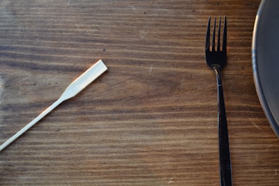 A wooden tabletop displays a single bamboo chopstick and a black fork. The dark fork is positioned near the edge of a round, dark-colored plate on the right, offering a contrast with the light wood background.