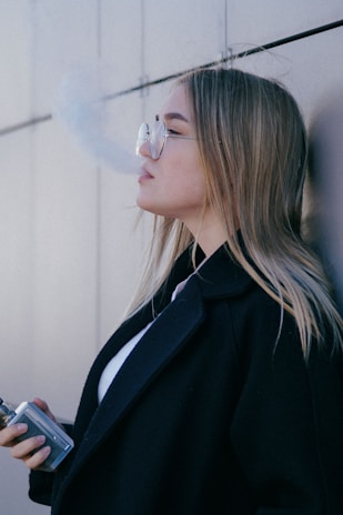 A person with long hair, wearing a black coat and glasses, is exhaling vapor against a neutral-colored wall. They are holding a vaping device in their hand, and the vapor creates a misty cloud extending from their mouth.