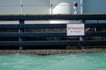A black, metallic barrier lines a waterfront, with a bold red and white 'No Trespassing No Tying Up' sign prominently displayed. Beneath the barrier, barnacles cling to the structure at the water's edge. The water below is a shade of turquoise, while a grey concrete wall or building provides the background with a red light fixture visible above the barrier.