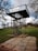 A metal observation platform with a staircase stands in the middle of a grassy park. The structure is elevated, supported by poles, and surrounded by trees in the background. A paved path leads up to the platform, and a few people are visible in the distance, walking on the grass.