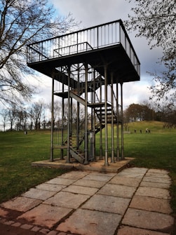 A metal observation platform with a staircase stands in the middle of a grassy park. The structure is elevated, supported by poles, and surrounded by trees in the background. A paved path leads up to the platform, and a few people are visible in the distance, walking on the grass.