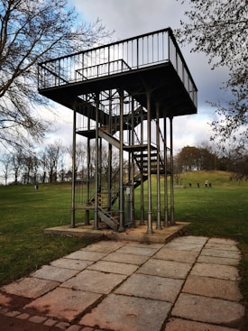 A metal observation platform with a staircase stands in the middle of a grassy park. The structure is elevated, supported by poles, and surrounded by trees in the background. A paved path leads up to the platform, and a few people are visible in the distance, walking on the grass.