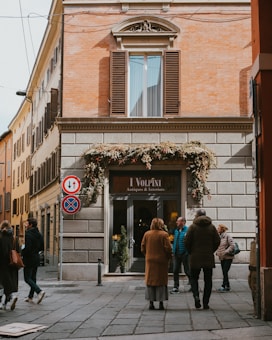 A street scene featuring a building with warm-toned bricks and a shop entrance labeled 'I Volpini Antiques & Interiors.' The facade is adorned with decorative foliage above the entrance. Several people are walking or standing near the shop, dressed in autumn or winter clothing.
