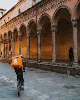 A person on a bicycle is riding along a cobblestone street under a series of arches in an old European-style building. The cyclist is carrying a large orange Just Eat delivery backpack. The building features ornate columns and aged, textured walls. Another person stands by a column in the shadows.