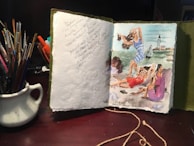 An open sketchbook on a table features a colorful painting on the right page depicting people relaxing on a beach with a lighthouse in the background. The left page contains handwritten notes. Several paintbrushes are held in a ceramic jug next to the book.