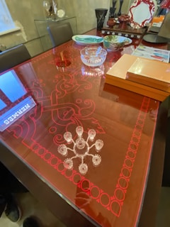 A modern dining table with a glossy red surface, intricately adorned with swirling patterns and bordered by a geometric design. Various elegant dishes and decorative items, including a pair of pepper mills, a glass bowl, and a stack of books or boxes, are arranged on the table. The background shows a minimalist setting with glass shelves displaying decorative pieces.