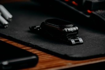A black smartwatch with a metal band is placed on a dark textured mat. The background includes hints of other tech items such as a pen and possibly a smartphone. The lighting is low, creating a sleek and modern atmosphere.