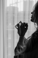 A silhouette of a person holding a small perfume bottle near the window. The room appears to be softly lit, with transparent curtains allowing light to filter through. The scene exudes a sense of mystery and calmness.