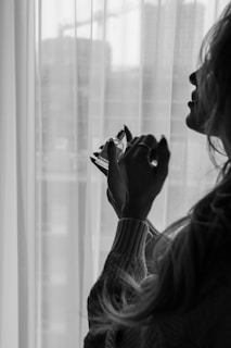 A silhouette of a person holding a small perfume bottle near the window. The room appears to be softly lit, with transparent curtains allowing light to filter through. The scene exudes a sense of mystery and calmness.