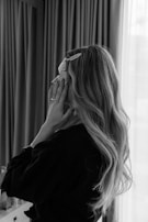 A person with long, wavy hair is standing indoors in front of a window with sheer curtains. They are touching their face and wearing a dark robe. There is a hair clip visible in their hair.