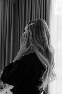 A person with long, wavy hair is standing indoors in front of a window with sheer curtains. They are touching their face and wearing a dark robe. There is a hair clip visible in their hair.