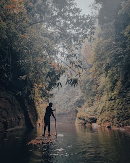 A person is navigating a bamboo raft through a serene river surrounded by lush, dense greenery and towering cliffs. The atmosphere is misty, creating a tranquil and slightly mysterious ambiance.
