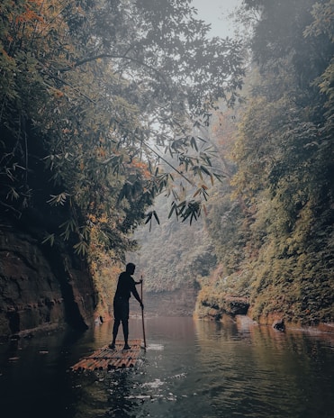 A person is navigating a bamboo raft through a serene river surrounded by lush, dense greenery and towering cliffs. The atmosphere is misty, creating a tranquil and slightly mysterious ambiance.