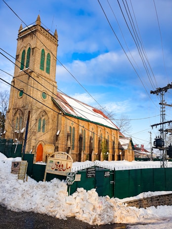 A large stone church with a tall tower stands surrounded by snow. The building features green-tinted windows and gothic architectural elements. Green fencing borders the perimeter, and there are several signs with text in a local script. Overhead, numerous power lines crisscross the sky under a clear blue with patches of clouds.