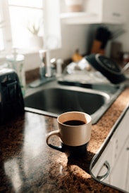 A warmly lit kitchen counter with a brown and beige speckled mug filled with coffee sits near a metal sink. Sunlight streams through a window, creating a bright, inviting atmosphere. In the background are kitchen items including a dish rack, some utensils, and a blurred potted plant.