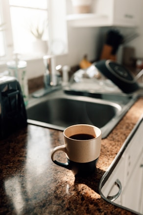 A warmly lit kitchen counter with a brown and beige speckled mug filled with coffee sits near a metal sink. Sunlight streams through a window, creating a bright, inviting atmosphere. In the background are kitchen items including a dish rack, some utensils, and a blurred potted plant.
