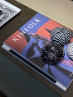 A magazine titled 'Kinfolk' is placed on a table with abstract artwork on the cover. Three glass spheres with black and white striped patterns rest on top. A second magazine partially enters the frame from the left.