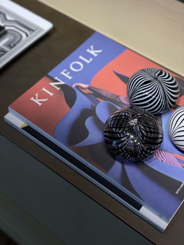 A magazine titled 'Kinfolk' is placed on a table with abstract artwork on the cover. Three glass spheres with black and white striped patterns rest on top. A second magazine partially enters the frame from the left.
