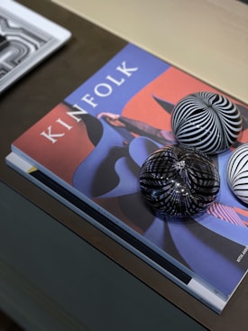 A magazine titled 'Kinfolk' is placed on a table with abstract artwork on the cover. Three glass spheres with black and white striped patterns rest on top. A second magazine partially enters the frame from the left.