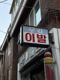 A traditional barber shop pole with a red and white spiral design is seen attached to a brick building. Above it, there's a sign with Korean text. The building features large windows and electrical wiring is visible along the walls.