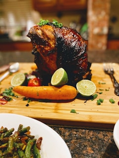 A roasted meat centerpiece garnished with fresh herbs sits on a wooden cutting board. Surrounding the meat are lime halves, a whole carrot, and scattered green herbs. In the foreground, a plate of seasoned green beans is visible.