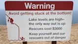 A warning sign on a sandy terrain close to a body of water, cautioning about the high lake levels and the difficulty of rescue, which is expensive. It advises people to avoid getting stuck and includes a graphic of a person struggling to climb out of a depression.