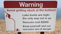A warning sign on a sandy terrain close to a body of water, cautioning about the high lake levels and the difficulty of rescue, which is expensive. It advises people to avoid getting stuck and includes a graphic of a person struggling to climb out of a depression.