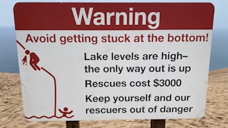 A warning sign on a sandy terrain close to a body of water, cautioning about the high lake levels and the difficulty of rescue, which is expensive. It advises people to avoid getting stuck and includes a graphic of a person struggling to climb out of a depression.