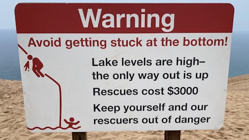 A warning sign on a sandy terrain close to a body of water, cautioning about the high lake levels and the difficulty of rescue, which is expensive. It advises people to avoid getting stuck and includes a graphic of a person struggling to climb out of a depression.