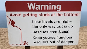 A warning sign on a sandy terrain close to a body of water, cautioning about the high lake levels and the difficulty of rescue, which is expensive. It advises people to avoid getting stuck and includes a graphic of a person struggling to climb out of a depression.