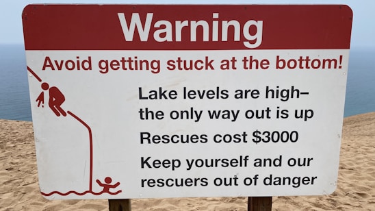 A warning sign on a sandy terrain close to a body of water, cautioning about the high lake levels and the difficulty of rescue, which is expensive. It advises people to avoid getting stuck and includes a graphic of a person struggling to climb out of a depression.