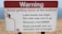 A warning sign on a sandy terrain close to a body of water, cautioning about the high lake levels and the difficulty of rescue, which is expensive. It advises people to avoid getting stuck and includes a graphic of a person struggling to climb out of a depression.