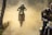 A motorcyclist is airborne, riding a dirt bike through a dusty terrain. The background is filled with clouds of dust, and another motorcyclist is visible on the right, partially obscured. The scene is set in an outdoor environment with trees visible on the side.