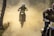 A motorcyclist is airborne, riding a dirt bike through a dusty terrain. The background is filled with clouds of dust, and another motorcyclist is visible on the right, partially obscured. The scene is set in an outdoor environment with trees visible on the side.