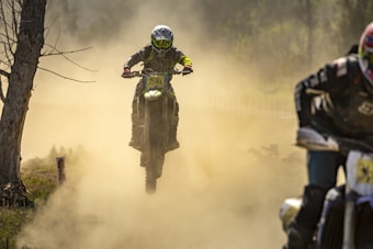 A motorcyclist is airborne, riding a dirt bike through a dusty terrain. The background is filled with clouds of dust, and another motorcyclist is visible on the right, partially obscured. The scene is set in an outdoor environment with trees visible on the side.