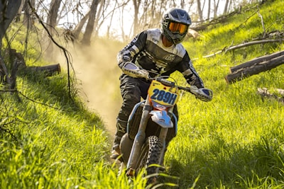A motorcyclist wearing a black and white outfit with a helmet rides through a dense, grassy forest path. Dust rises behind the bike, suggesting motion and speed. Sunlight filters through the trees, casting shadows and highlighting the vibrant green of the grass.