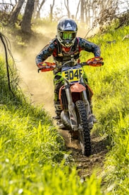 A motorcyclist wearing a helmet and goggles is navigating a dirt trail through a lush, green forest. Dust is rising from the path as the motorcycle speeds ahead. The rider wears a colorful racing suit and the front of the bike displays the number 304F.