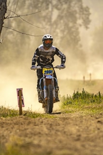 A motocross rider wearing protective gear and a helmet rides a dirt bike along a dusty track. The background is blurred, emphasizing the speed and focus on the rider, and there are sparse trees and vegetation visible along the path.