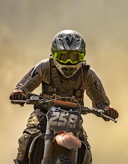 A motorcycle rider is captured in action, covered in dirt and dust. The individual wears protective gear including a helmet with a distinct pattern and bright yellow goggles. The number 258 is prominently displayed on the front of the motorcycle, which appears rugged and worn from use.