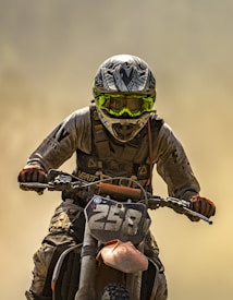 A motorcycle rider is captured in action, covered in dirt and dust. The individual wears protective gear including a helmet with a distinct pattern and bright yellow goggles. The number 258 is prominently displayed on the front of the motorcycle, which appears rugged and worn from use.