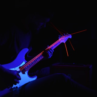 A musician playing an electric guitar is partially illuminated in blue and red light, creating a dramatic and moody atmosphere. Bright red laser-like lines trace along the strings and headstock, adding a dynamic visual effect.