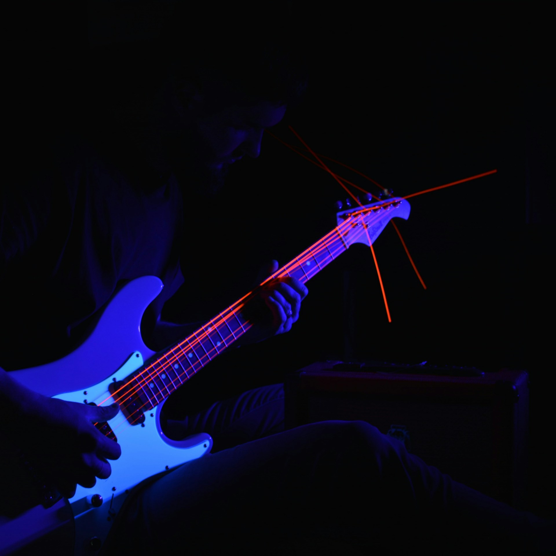A musician playing an electric guitar is partially illuminated in blue and red light, creating a dramatic and moody atmosphere. Bright red laser-like lines trace along the strings and headstock, adding a dynamic visual effect.