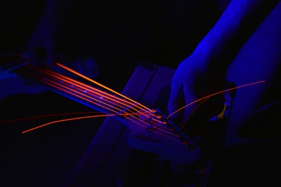 A close-up view of a person's hands adjusting the tuning pegs of a guitar with neon orange strings against a dark, dimly lit background. The strings glow brightly, contrasting with the blue ambient lighting that casts shadows and highlights over the textured surface of the guitar body.