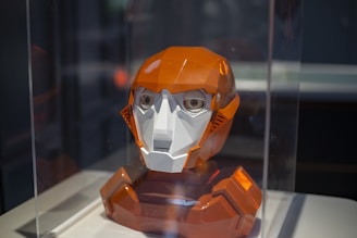 A geometric robotic head displayed inside a transparent protective case. The head features a combination of orange and white colors with a glossy finish and detailed facial features, including large eyes and angular structures. It is positioned on a pedestal within a dimly lit setting.