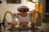 A humanoid robot with white and orange coloring is positioned inside a glass enclosure. The robot has a rounded head with a dark visor and red-lit eyes, and its arms are connected to machinery with flexible tubing. The background includes blurred interior elements with warm lighting.